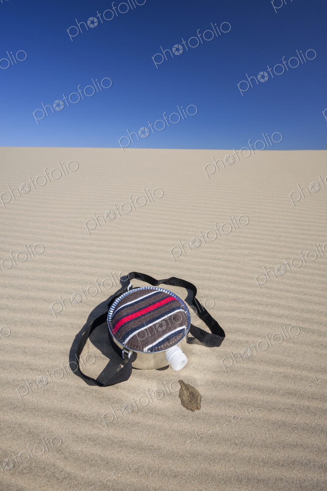A fabric-covered canteen rests on rippled sand dunes with a small puddle of spilled…