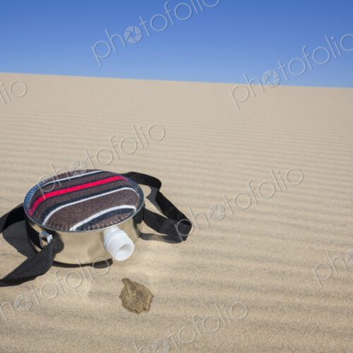 A fabric-covered canteen rests on rippled sand dunes with a small puddle of spilled…