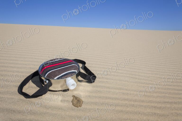 A fabric-covered canteen rests on rippled sand dunes with a small puddle of spilled…