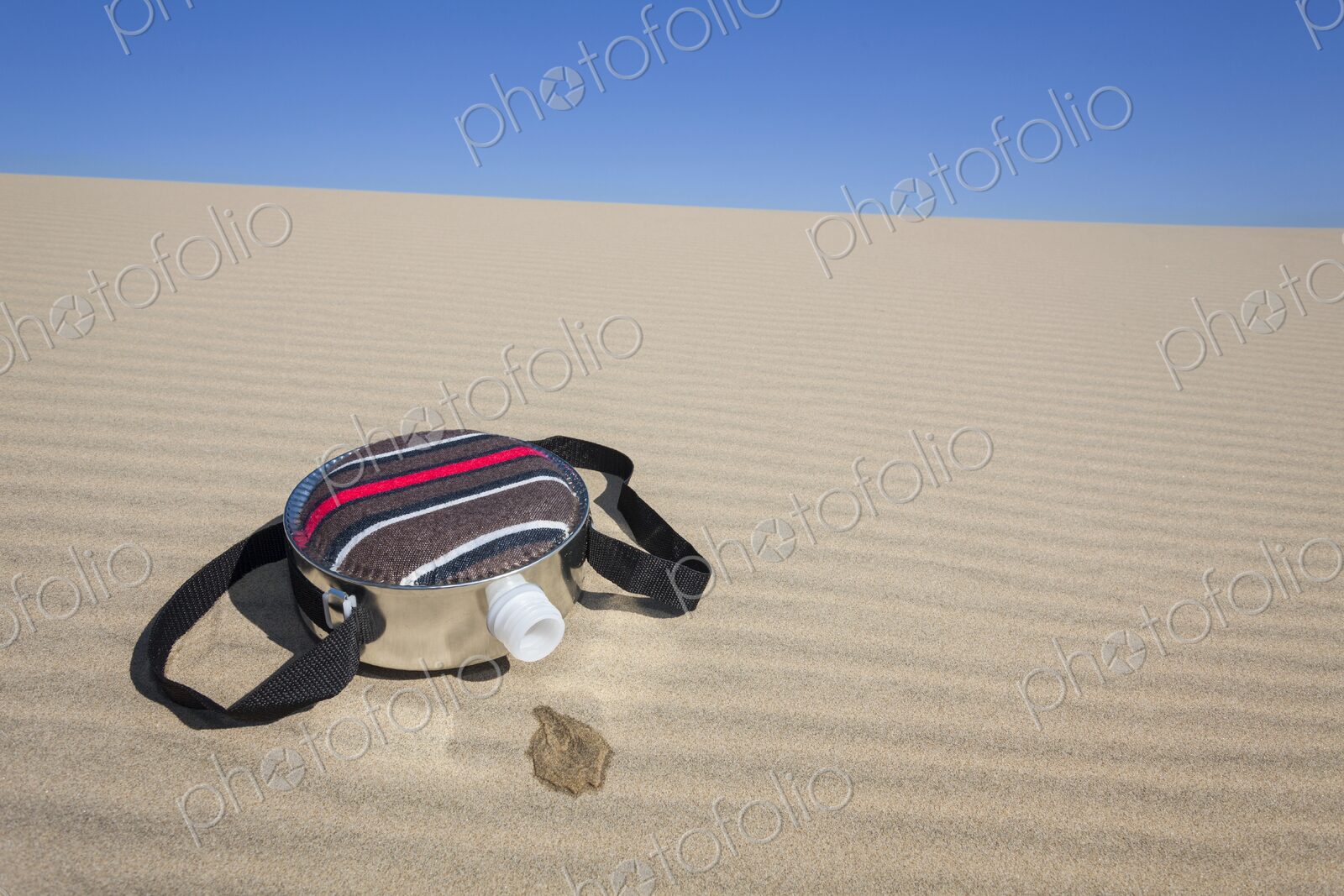 A fabric-covered canteen rests on rippled sand dunes with a small puddle of spilled…