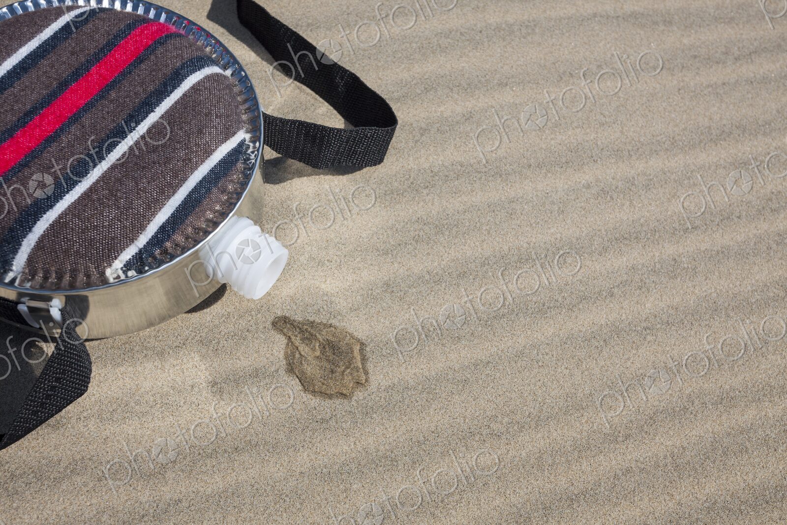 A fabric-covered canteen rests on rippled sand dunes with a small puddle of spilled…