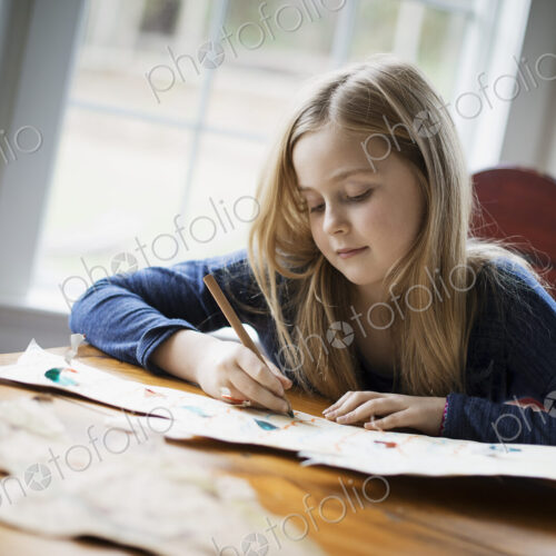 A family home. A young girl sitting at a table drawing on a large piece of paper.…