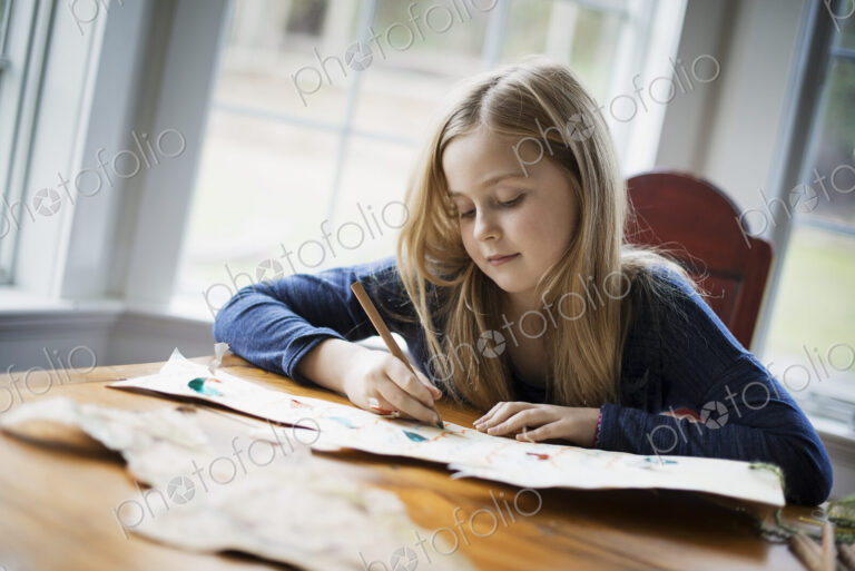 A family home. A young girl sitting at a table drawing on a large piece of paper.…