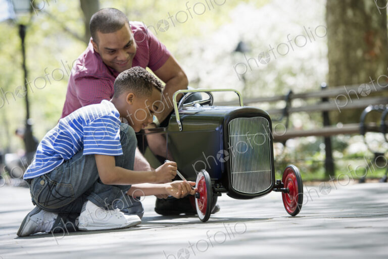 A family in the park on a sunny day. A boy repairing an old fashioned toy peddle car.