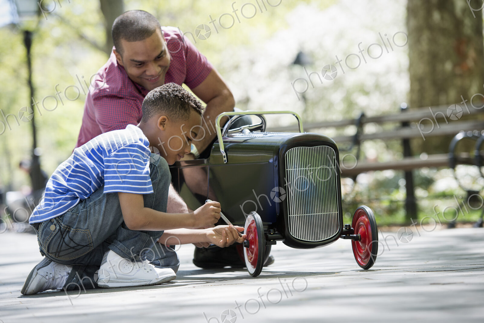 A family in the park on a sunny day. A boy repairing an old fashioned toy peddle car.