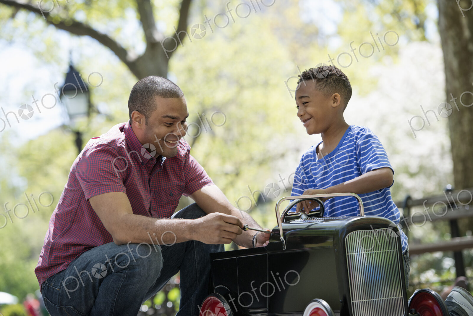 A family in the park on a sunny day. A boy riding an old fashioned toy peddle car.