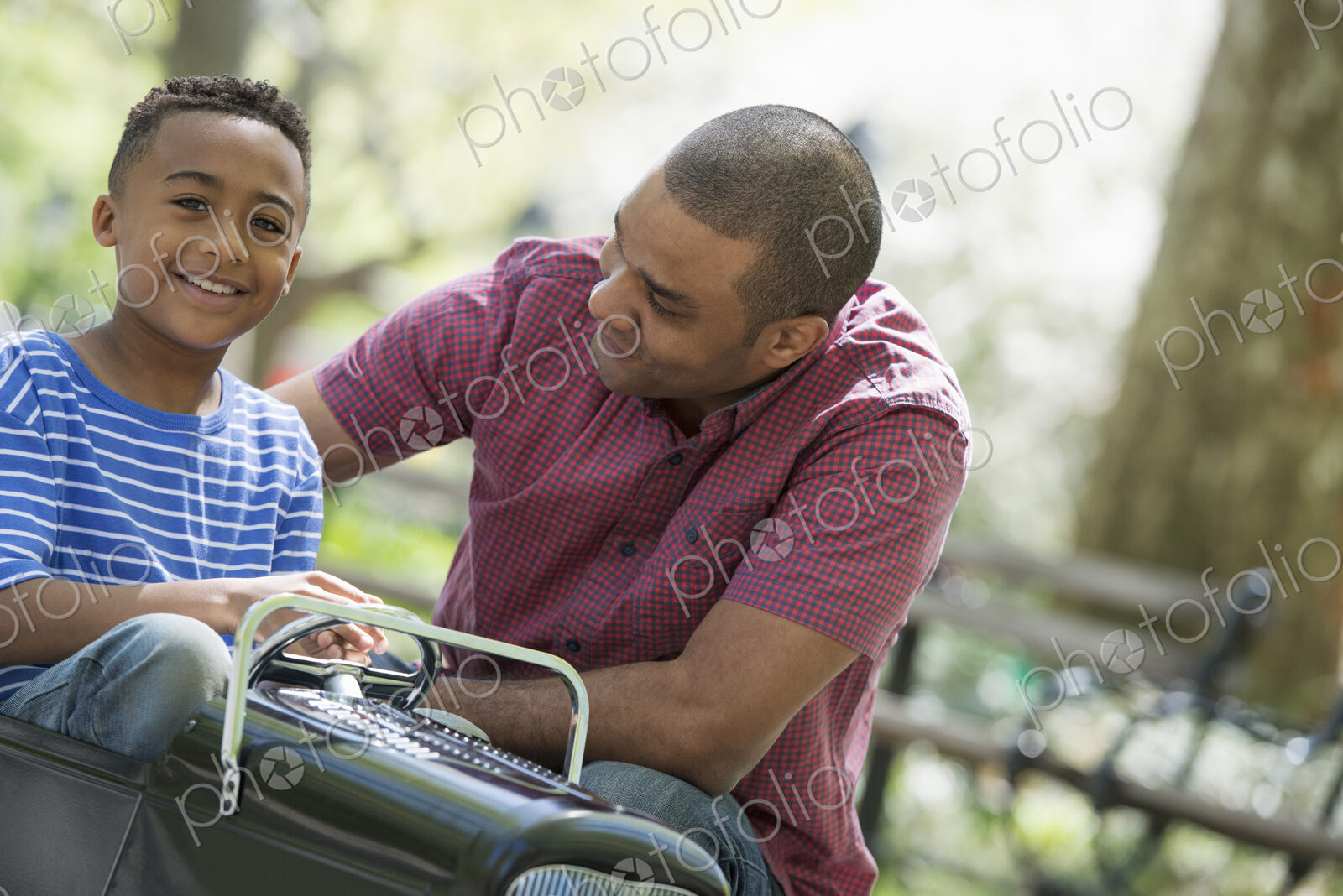 A family in the park on a sunny day. A boy riding an old fashioned toy peddle car.