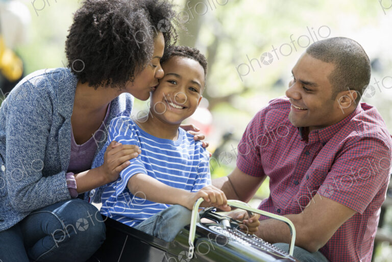 A family in the park on a sunny day. A boy riding an old fashioned toy peddle car.