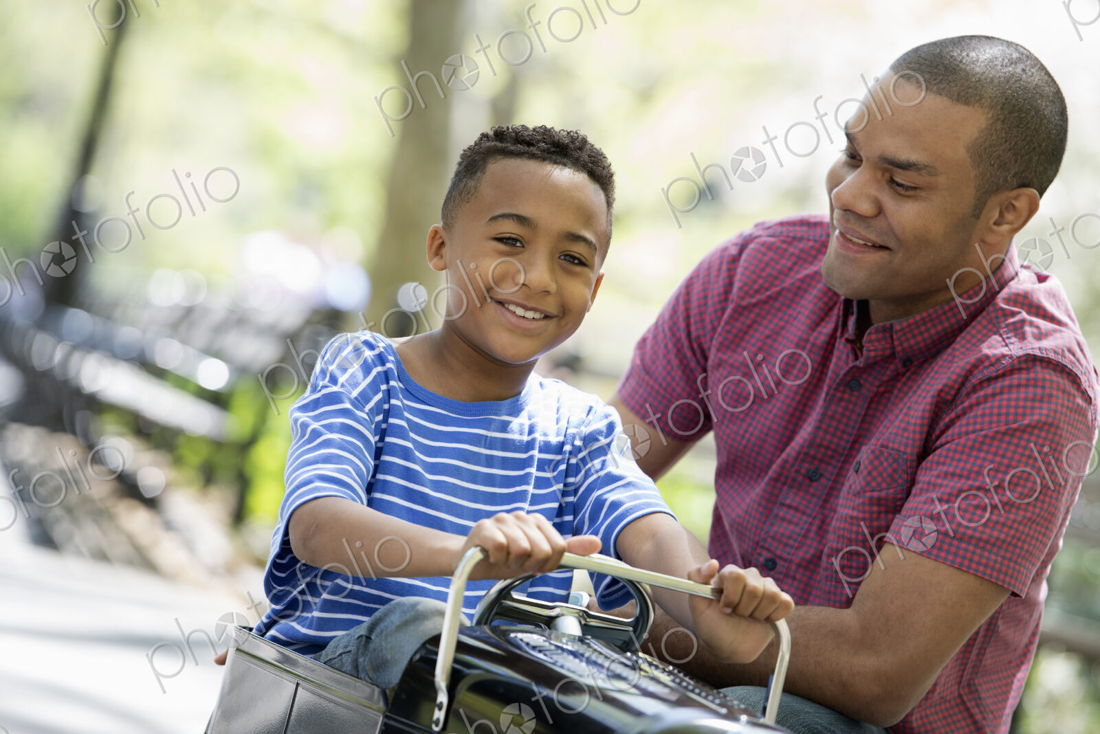 A family in the park on a sunny day. A boy riding an old fashioned toy peddle car.