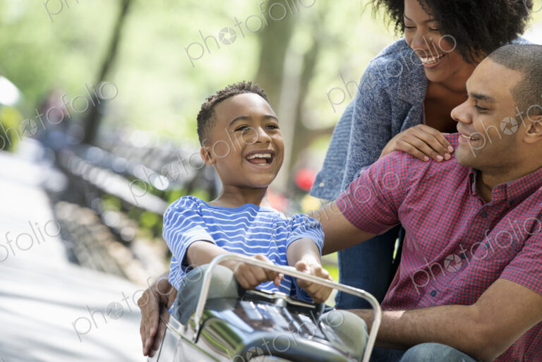 A family in the park on a sunny day. A boy riding an old fashioned toy peddle car.