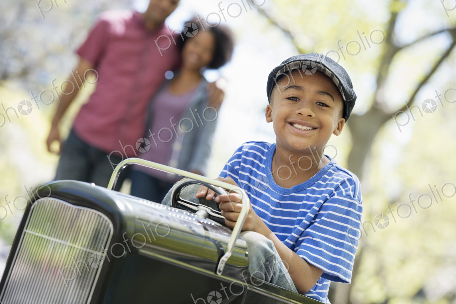 A family in the park on a sunny day. A boy riding an old fashioned toy peddle car.