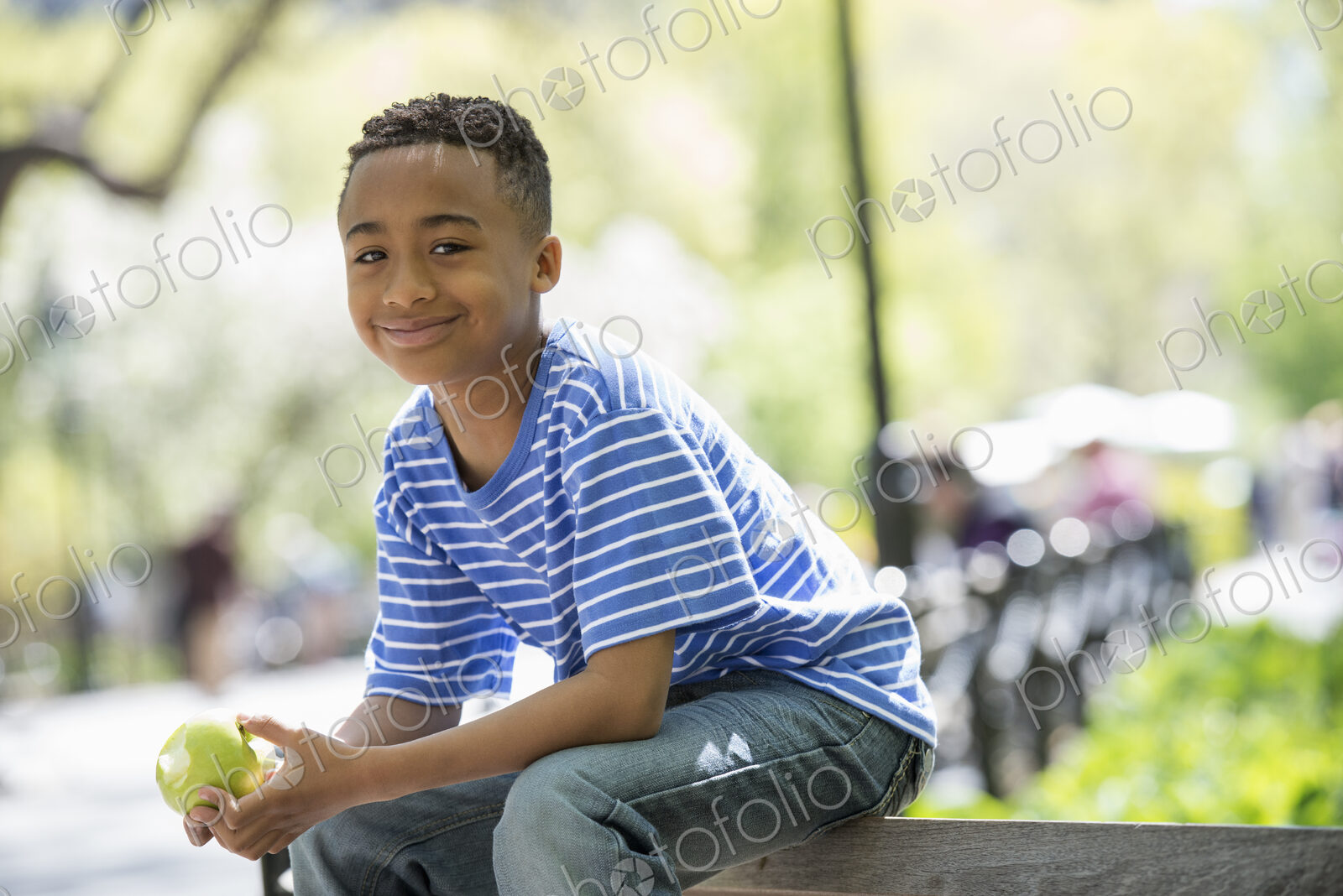 A family in the park on a sunny day. A boy sitting on a fence eating an apple.