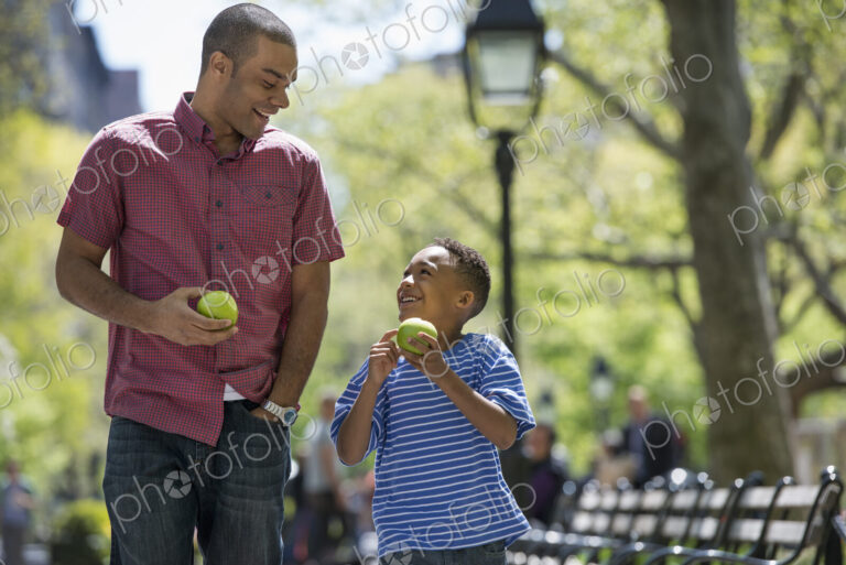 A family in the park on a sunny day. A man and a boy eating apples.