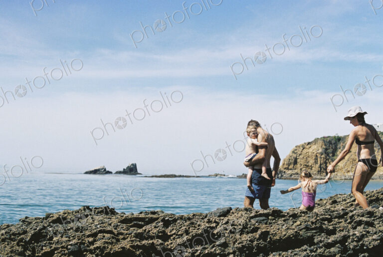 A family on holiday, two adults with their son and daughter walking across rocks by…