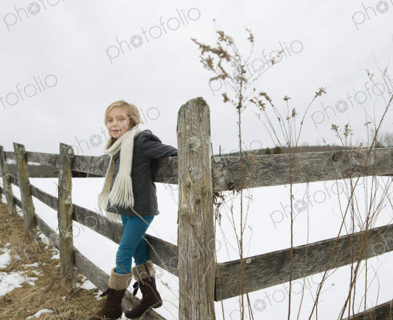 A farm in the snow. A girl with a hat and scarf and boots, leaning on a fence.