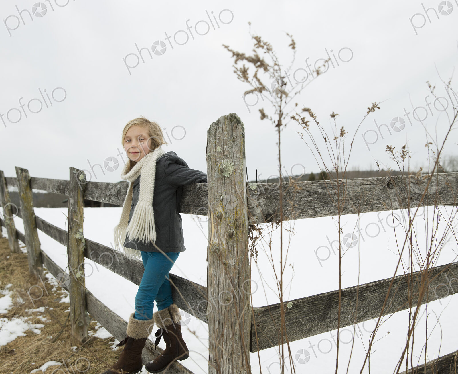 A farm in the snow. A girl with a hat and scarf and boots, leaning on a fence.