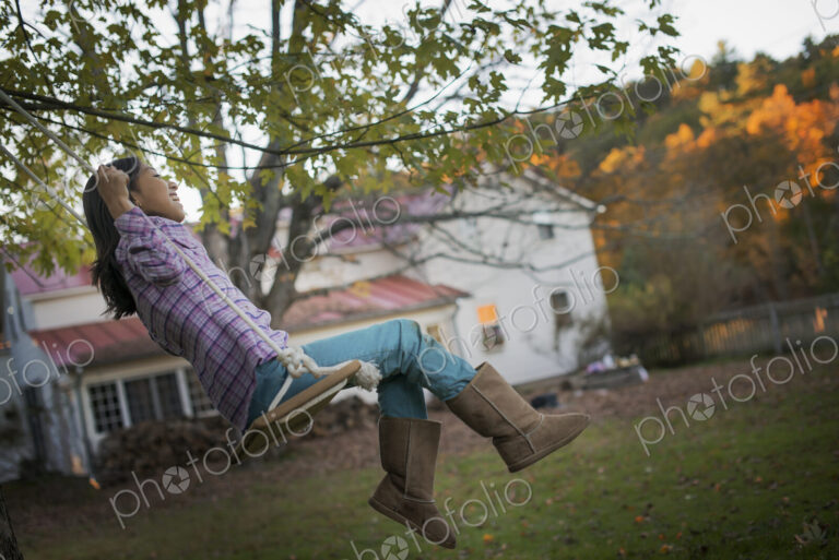 A farmhouse garden, Autumn trees lit by late sunshine. A child seated on a swing.