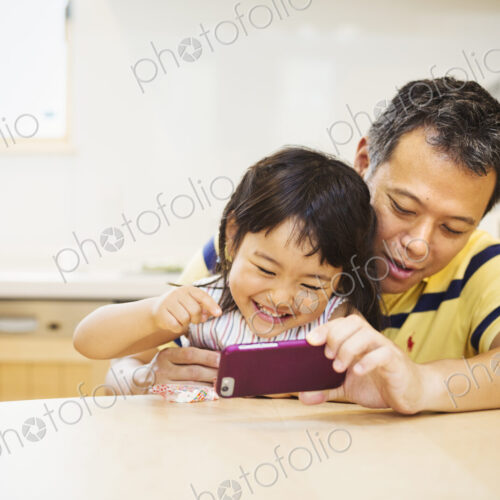 A father and daughter sitting at a table playing with a smart phone, looking at the…