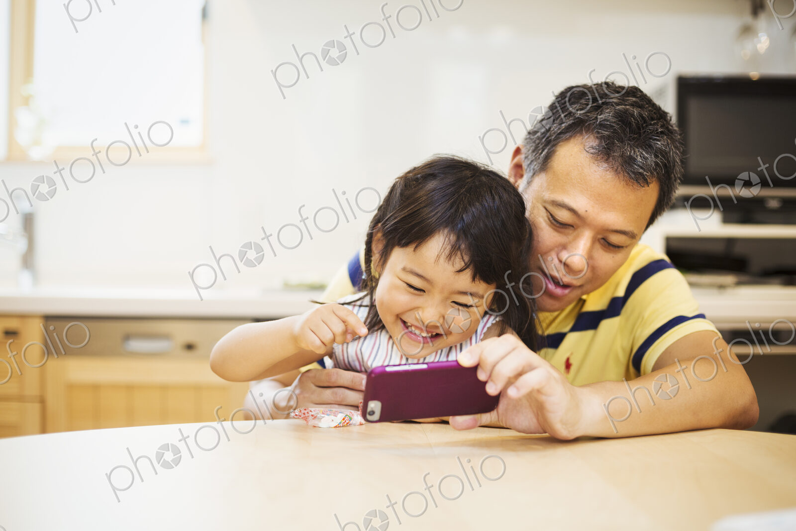 A father and daughter sitting at a table playing with a smart phone, looking at the…
