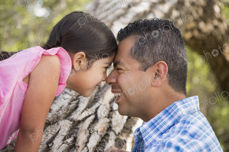 A father and daughter with foreheads pressed together by a tree in the park.