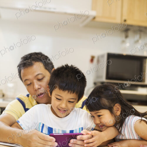 A father, son and daughter sitting at a table playing with a smart phone, looking at…