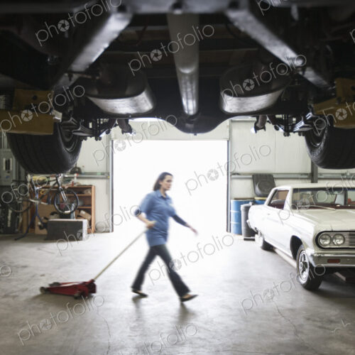 A female car mechanic pulls a portable car jack acrosss the floor of the car repair shop.