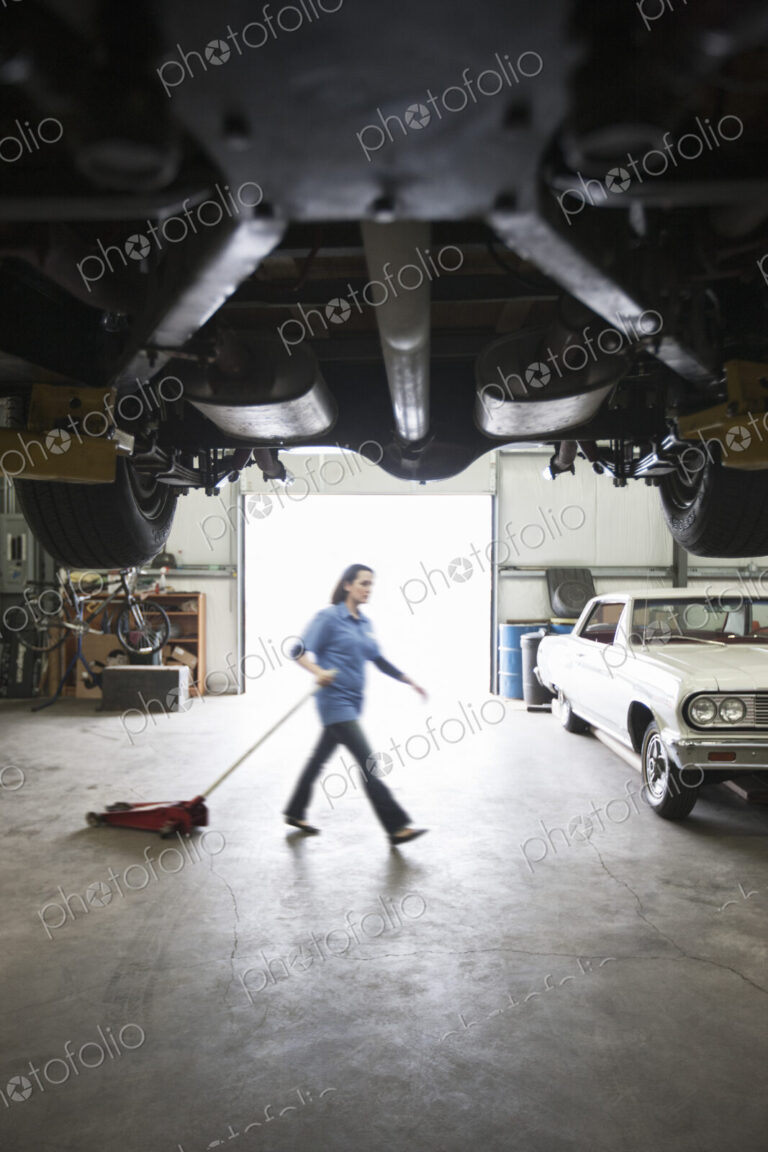 A female car mechanic pulls a portable car jack acrosss the floor of the car repair shop.