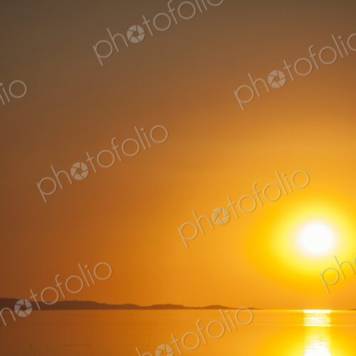 A fiery orange sun sets over the calm surface of the Great Salt Lake, casting golden…