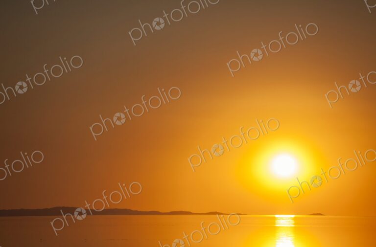 A fiery orange sun sets over the calm surface of the Great Salt Lake, casting golden…