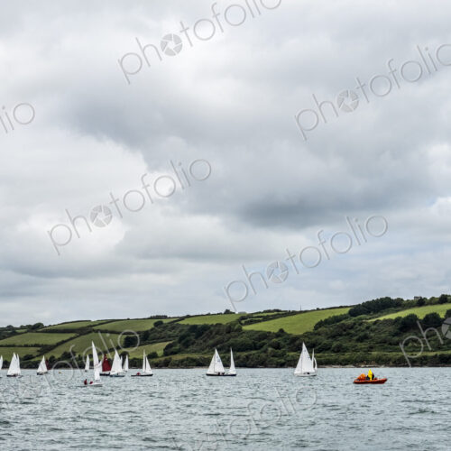 A fleet of sailing boats on the water offshore with a larger yacht and a safety…