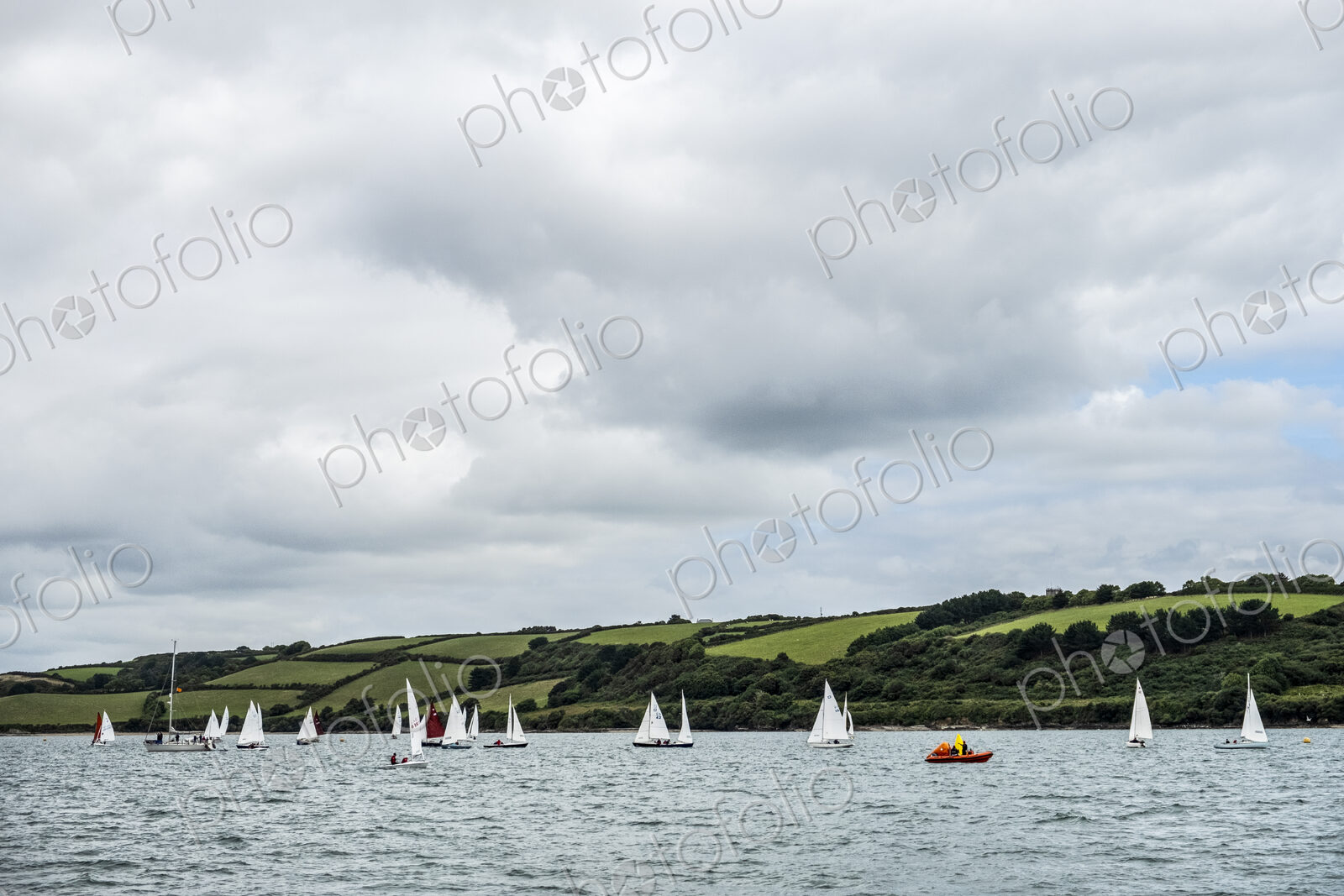 A fleet of sailing boats on the water offshore with a larger yacht and a safety…