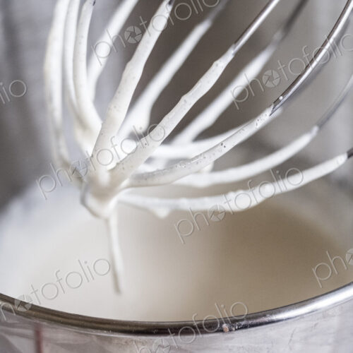 A food mixer whisk coated with batter above a metal bowl with ingredients. A food mixer whisk coated with batter above a metal bowl with ingredients.