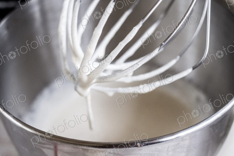 A food mixer whisk coated with batter above a metal bowl with ingredients.