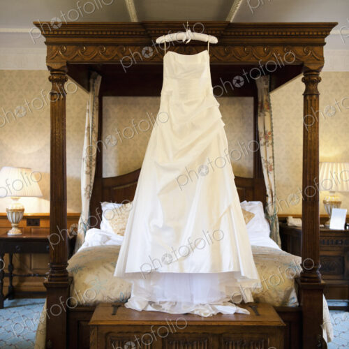 A full length white wedding dress hanging from a hanger on a four-poster bed. A full length white wedding dress hanging from a hanger on a four-poster bed.