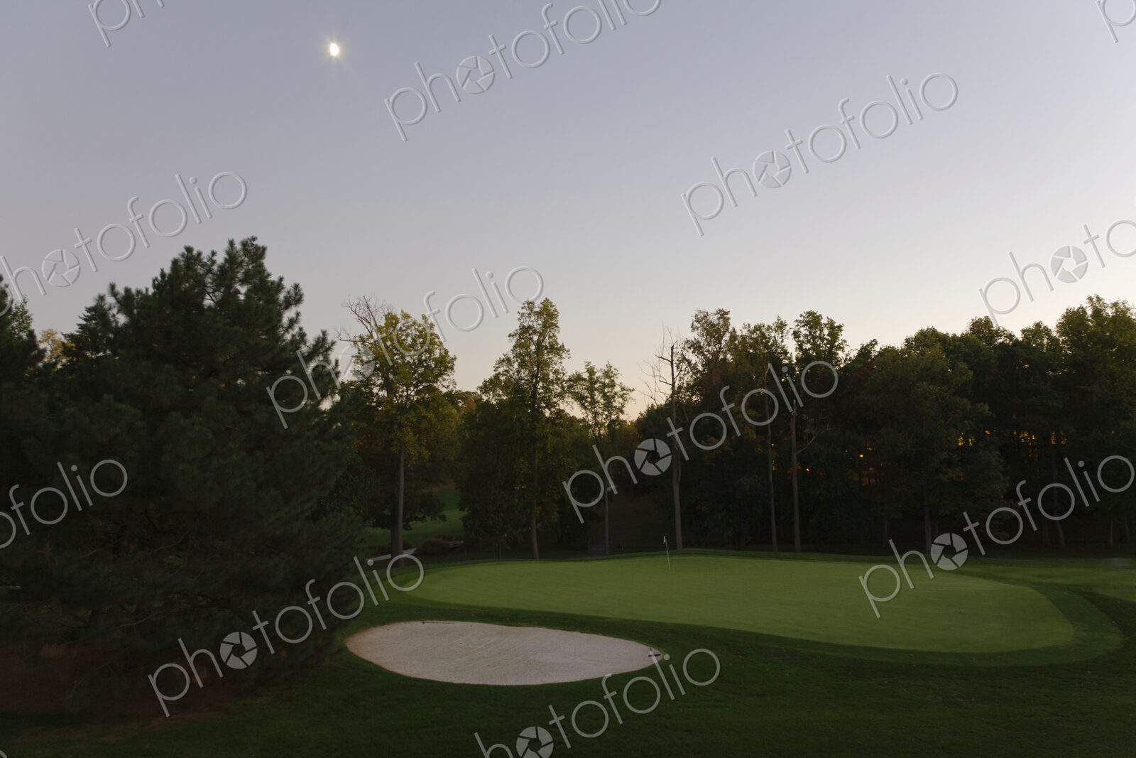 A full moon in the sky at dusk, hanging over a sand trap on a golf course.