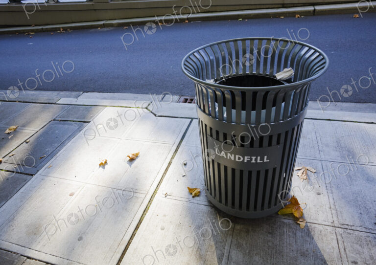 A garbage can on the sidewalk in the city.