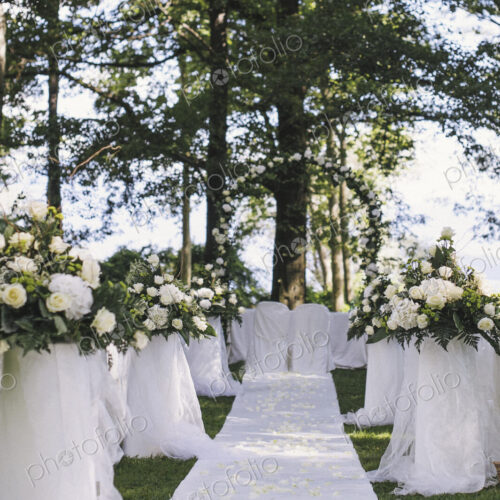 A garden with tables laid under the shade of tall trees, a floral arch, setting for a…