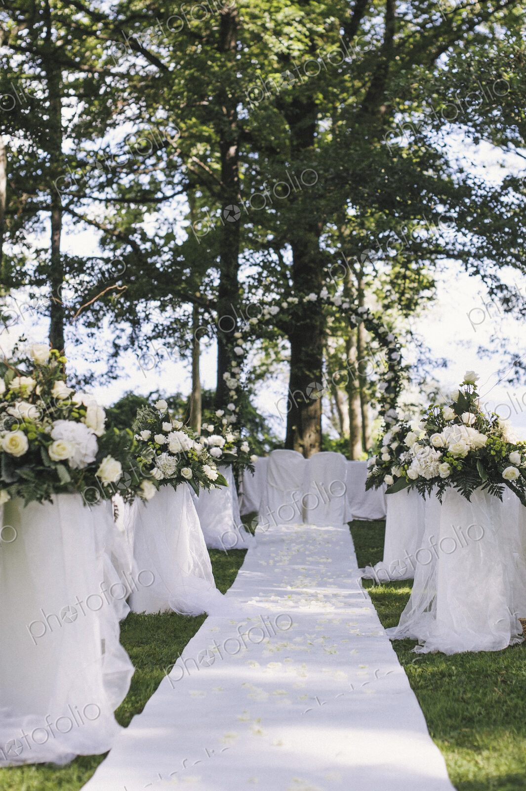 A garden with tables laid under the shade of tall trees, a floral arch, setting for a…
