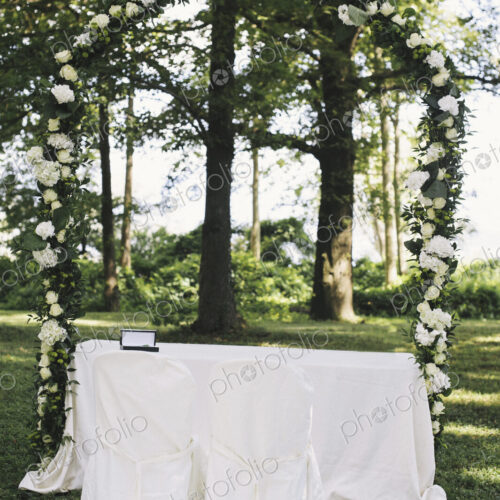 A garden with tables laid under the shade of tall trees, a floral arch, setting for a… A garden with tables laid under the shade of tall trees, a floral arch, setting for a…