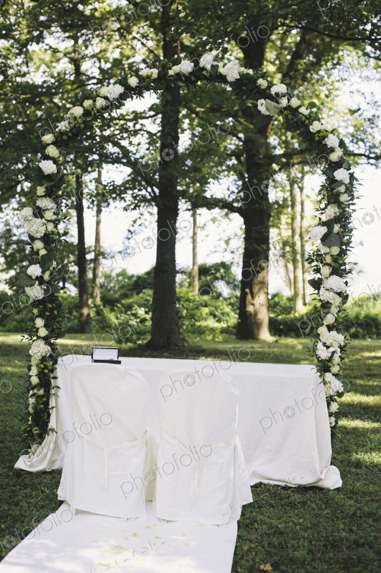 A garden with tables laid under the shade of tall trees, a floral arch, setting for a…