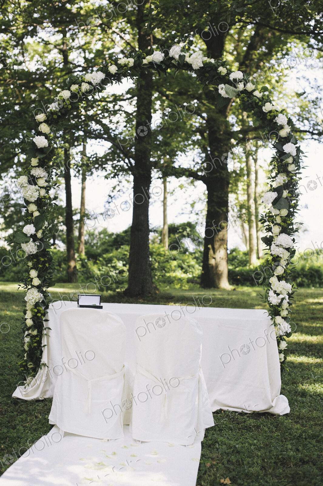 A garden with tables laid under the shade of tall trees, a floral arch, setting for a…