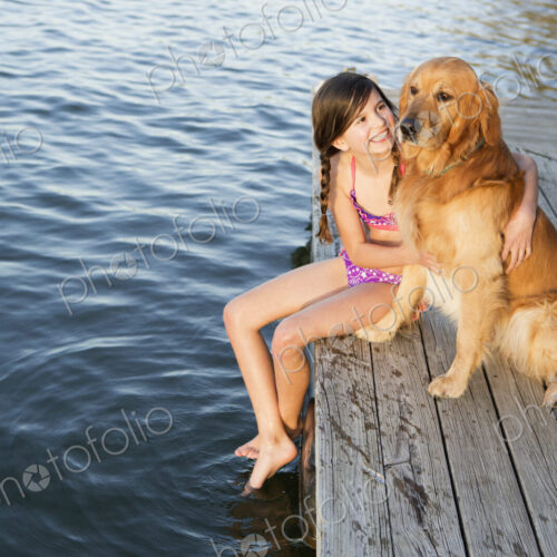 A girl and her golden retriever dog seated on a jetty by a lake.