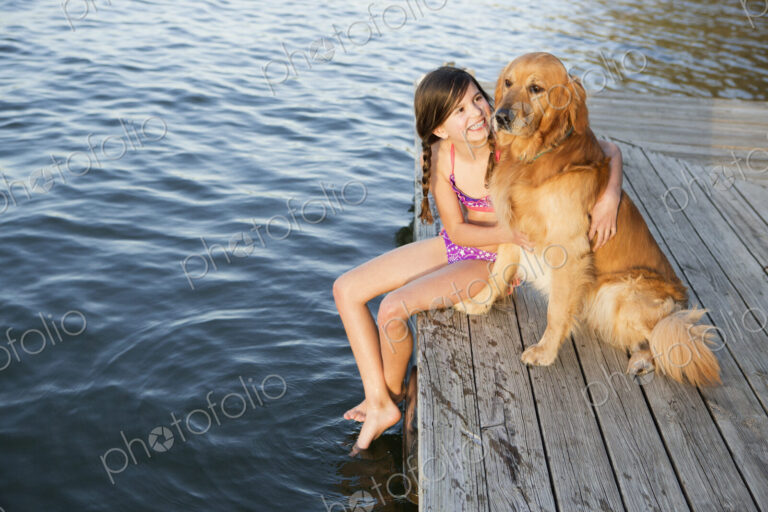 A girl and her golden retriever dog seated on a jetty by a lake.