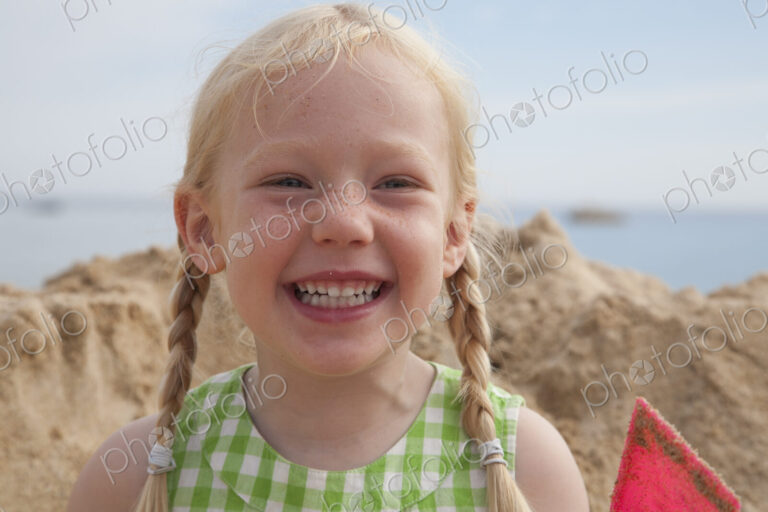 A girl beside a huge pile of sand on the beach, grinning at the camera.