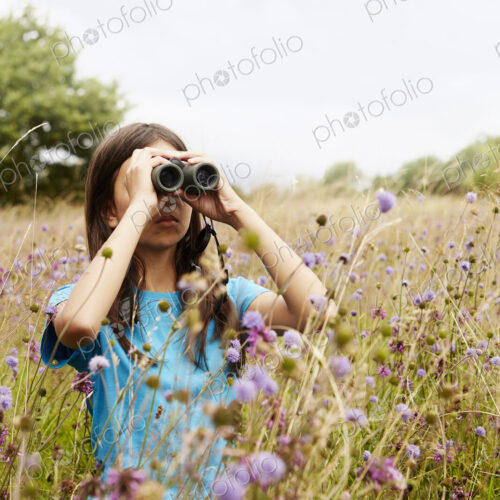 A girl holding binoculars, a young bird watcher standing in a meadow of tall grass and… A girl holding binoculars, a young bird watcher standing in a meadow of tall grass and…