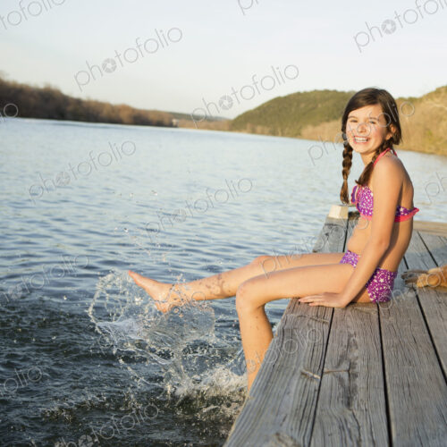 A girl in a bikini sitting on a jetty with her feet in the water.