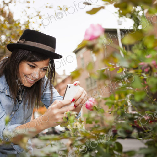 A girl in a brown trilby hat, taking a picture of roses with a smart phone.