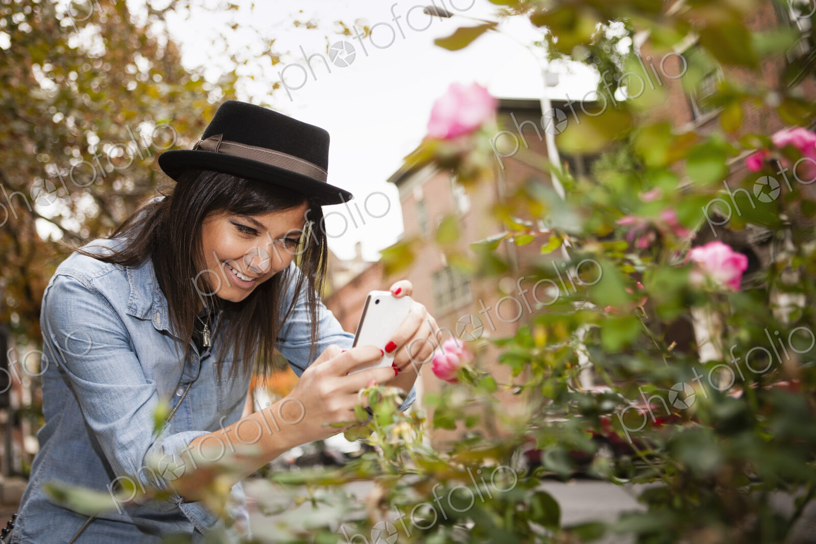 A girl in a brown trilby hat, taking a picture of roses with a smart phone.