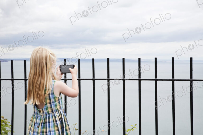 A girl in a checked sundress taking a photograph with a smart phone.