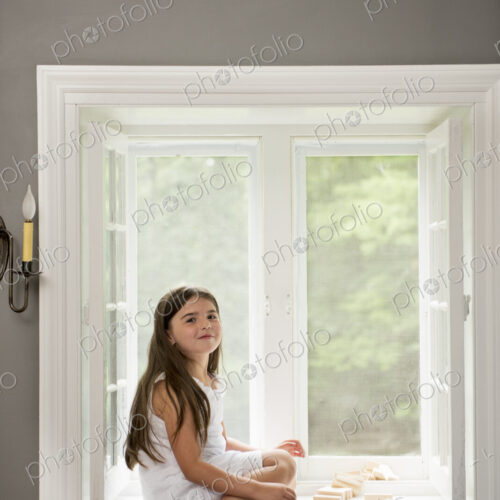 A girl in a white dress seated by a window playing.